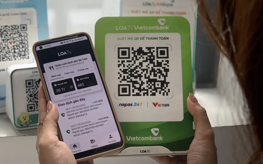 Chủ cửa hàng cầm điện thoại mở app Loa X cạnh bảng mã QR thanh toán của loa Loa X